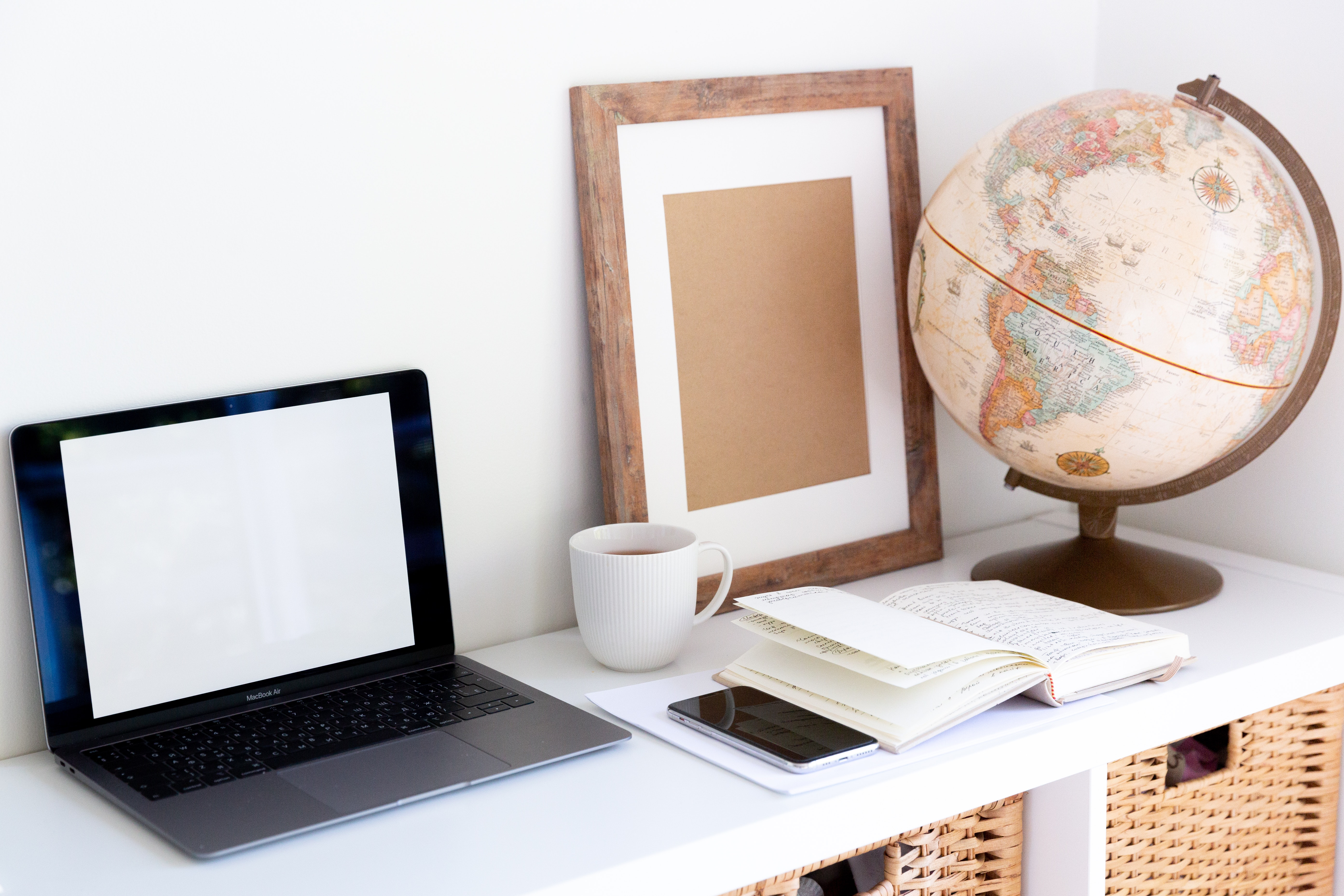 An open laptop, empty picture frame, book, phone, mug, and globe on a white shelf.