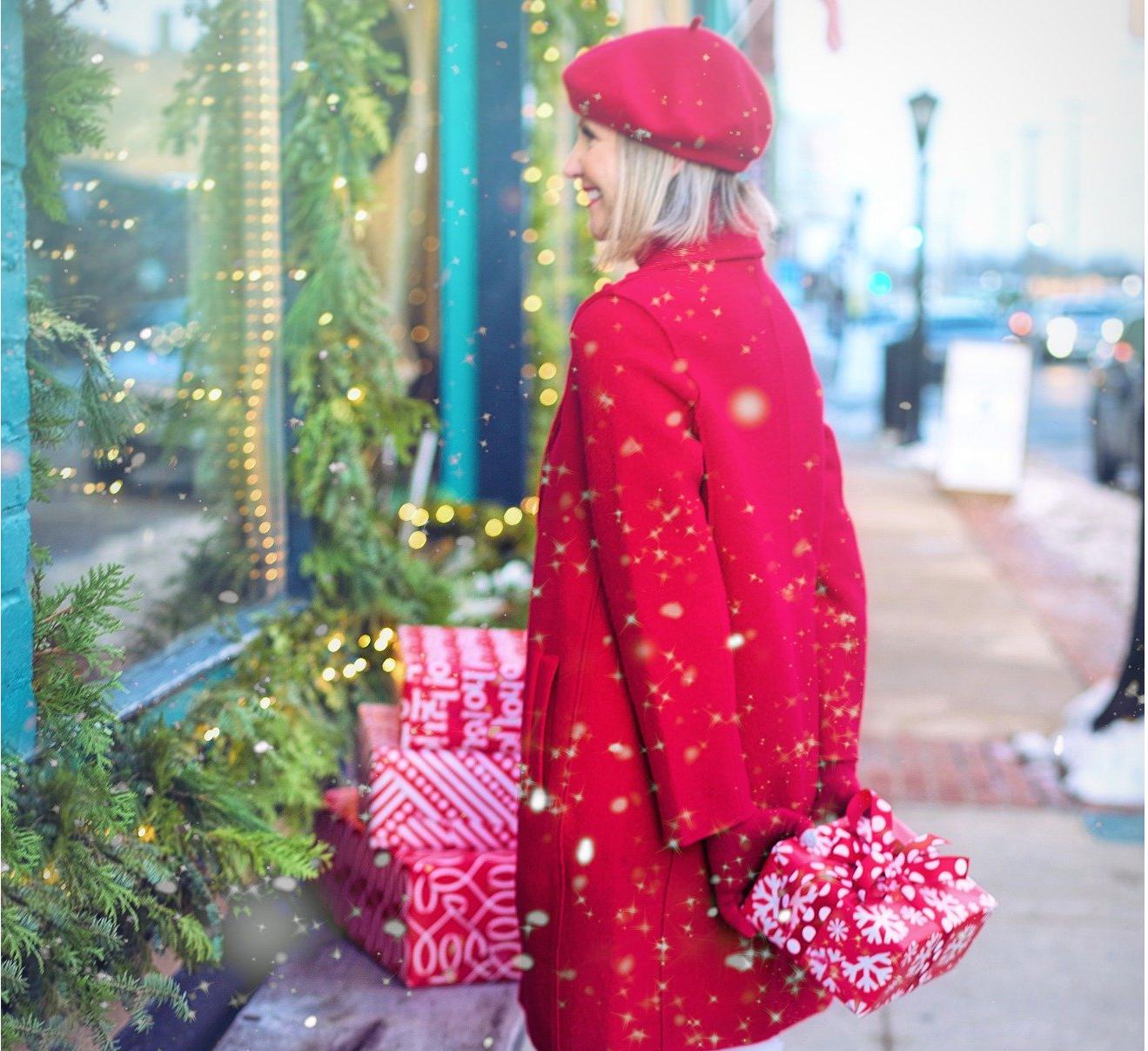 Woman in a red jacket and hat holding a wrapped present behind her back. She is outside on a snowy day looking in a store window that is decorated for the holidays.
