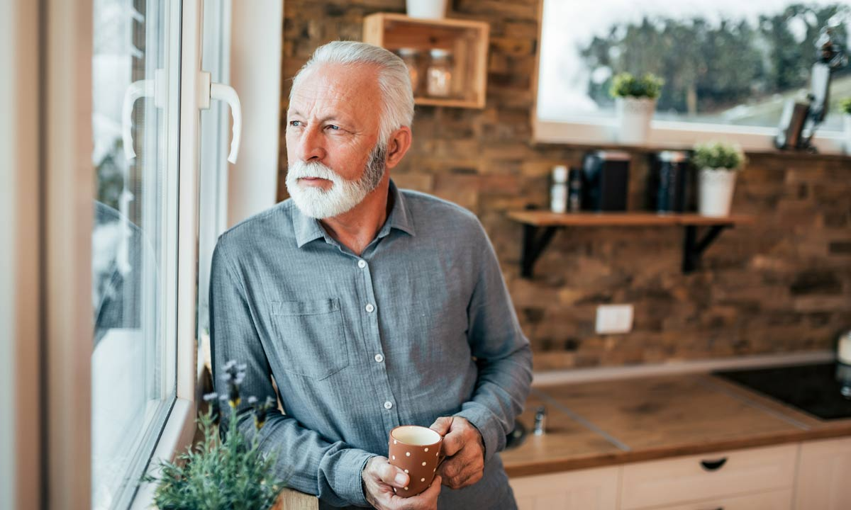 Elderly man in a grey shirt holding a coffee cup. He is in a kitchen and looking out the window.