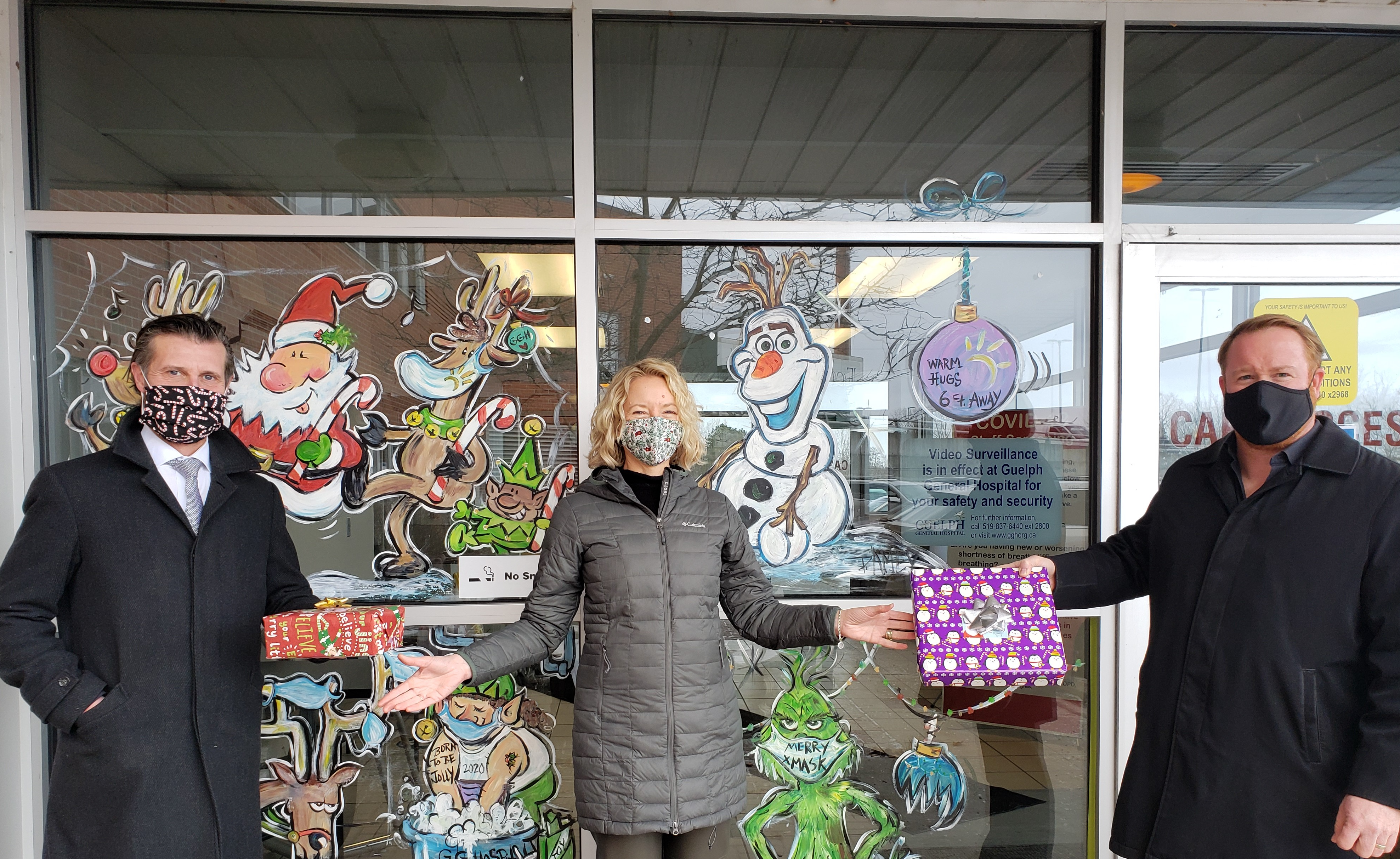 Two men and one woman standing in front of a holiday decorated window. The men are handing the woman wrapped presents.