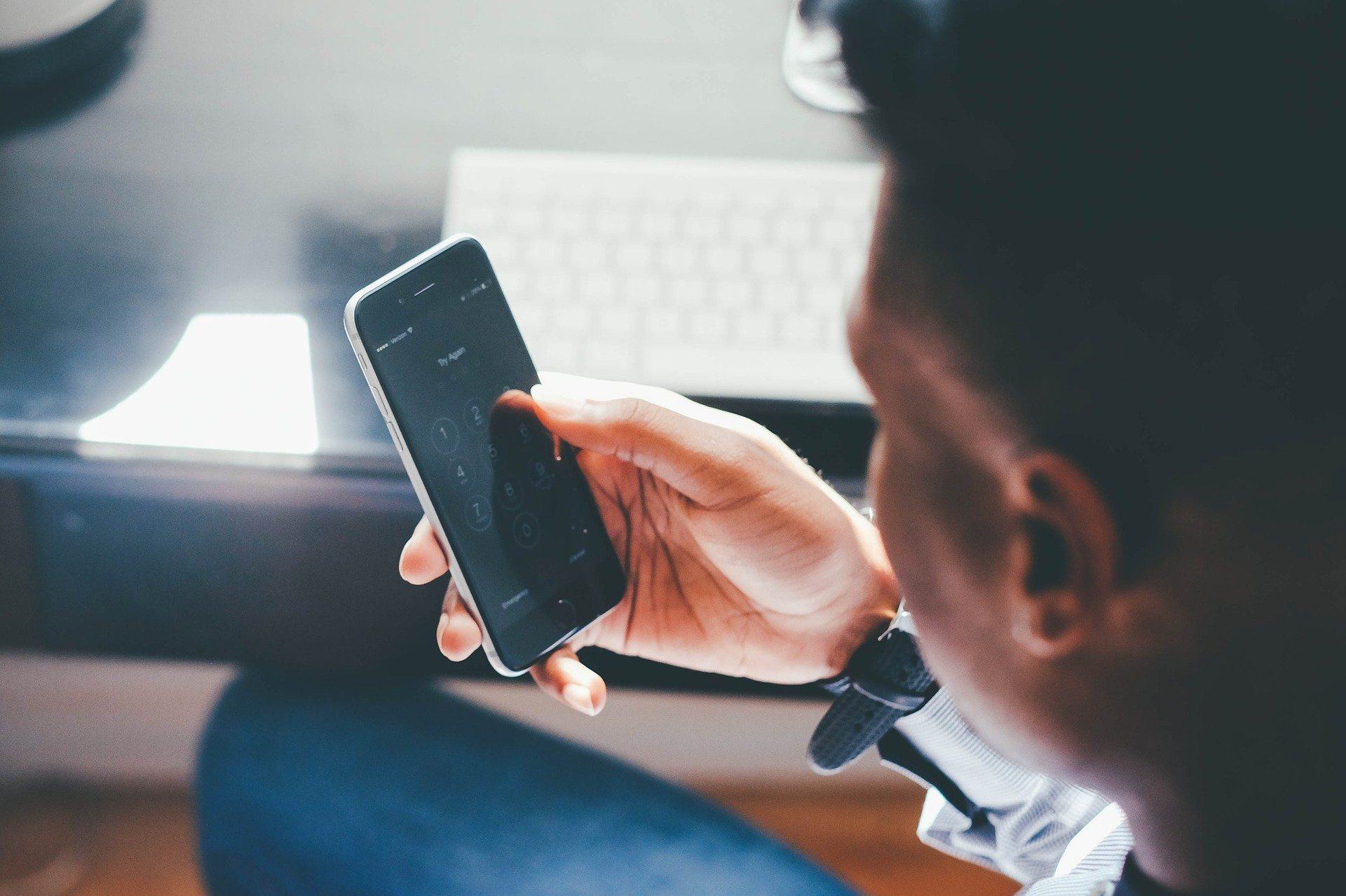 Man in jeans holding a mobile device. There is a desk and keyboard in the background.