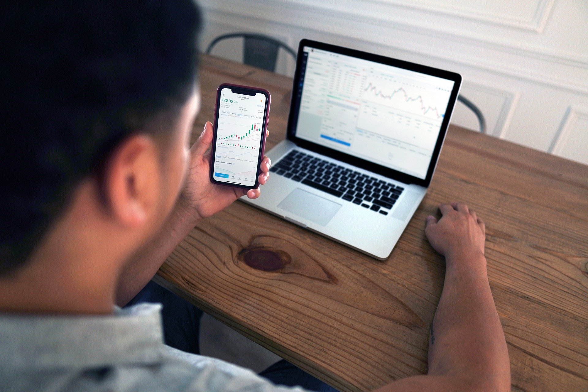 Man holding a mobile device, sitting a desk with an open laptop. Both screens are displaying graphs.