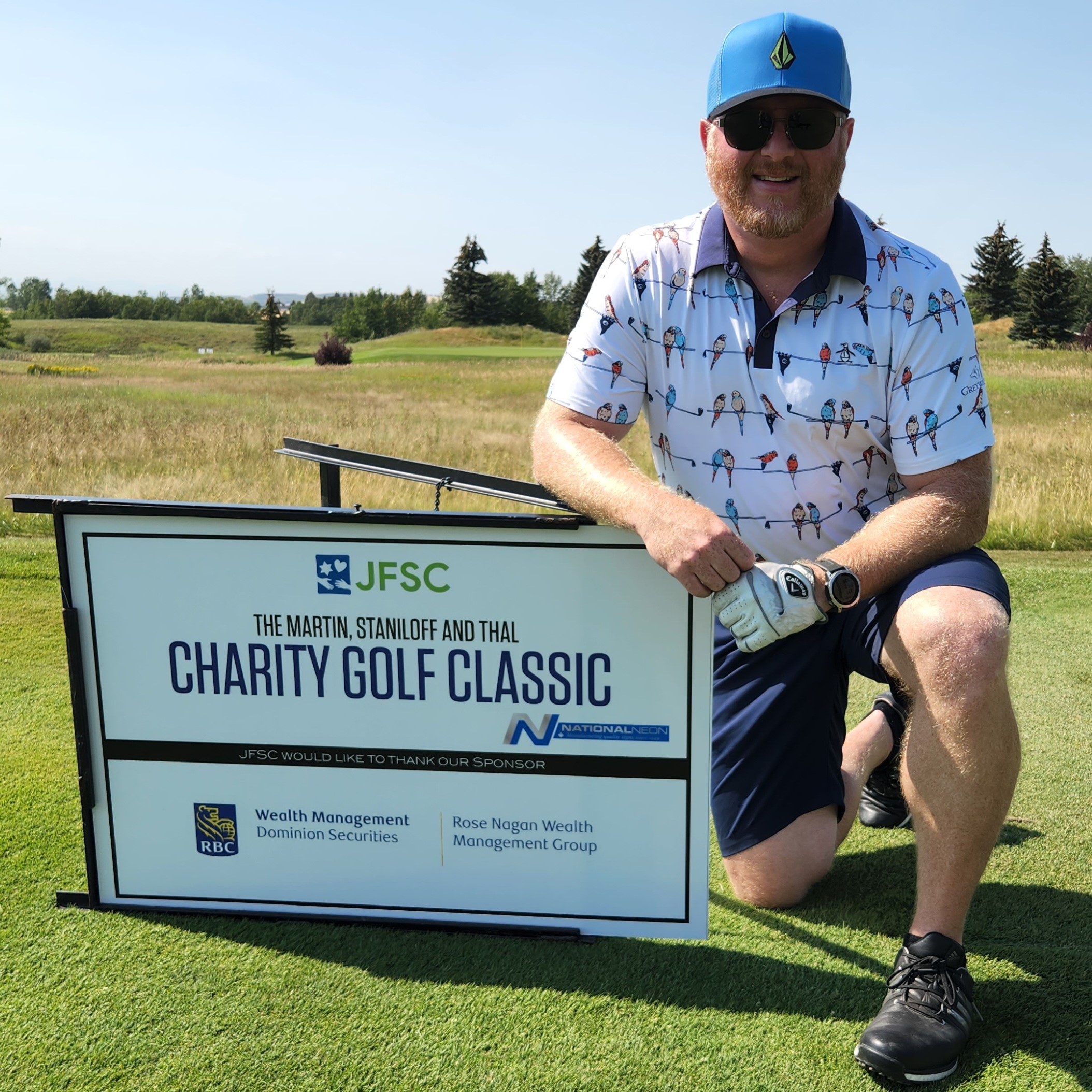 smiling man wearing golf gear kneeling next to a sign that says charity golf classic
