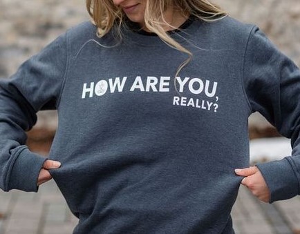Woman wearing a grey sweatshirt that reads "How are you, really?" in white text. She is standing outside with a blurred background. 
