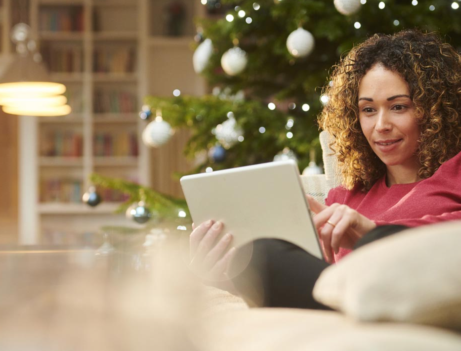 Woman sitting on a couch looking at her tablet. She is sitting in front of a Christmas tree. 