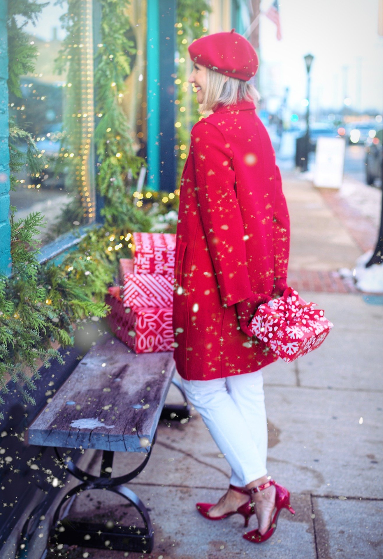 Woman in a red jacket and hat holding a wrapped present. She is standing outside in the snow looking in a store window that is decorated in greenery. 