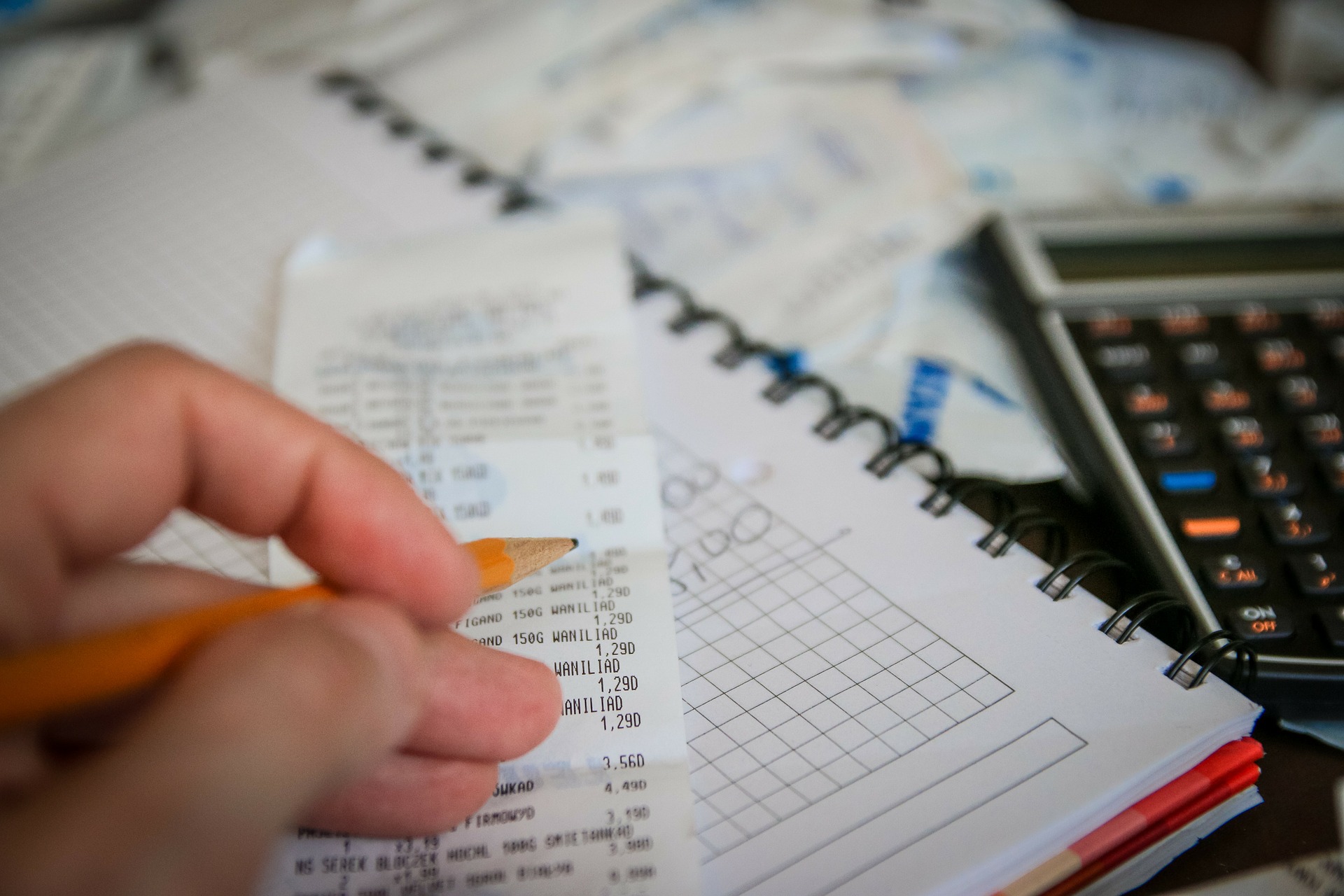 A close-up picture of receipts, notebook, and calculator on a tabletop. There is a hand holding a pencil above one of the receipts. 