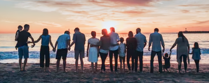 A multigenerational family holding hands on the beach as they look out at the sunset. 
