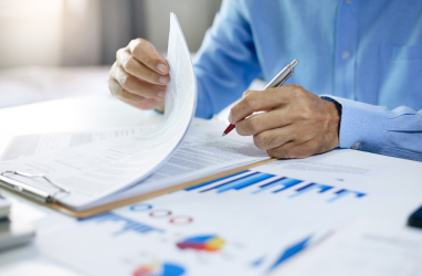 Man writing on one of several documents splayed in front of him on a table