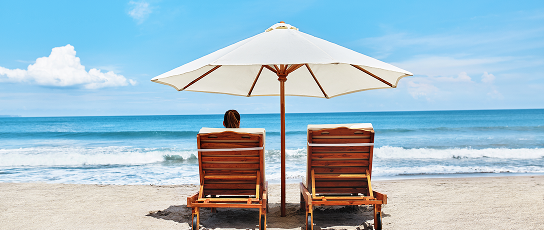 Person sitting on one of two lawn chairs under an umbrella at the beach.