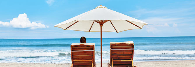 Person sitting on one of two lawn chairs under an umbrella at the beach.