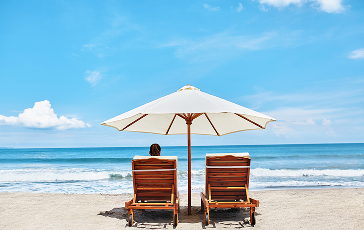 Person sitting on one of two lawn chairs under an umbrella at the beach.