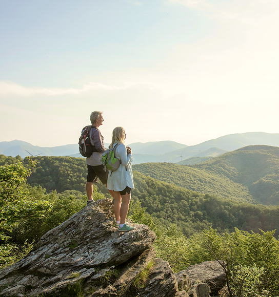 A couple on the summit of a hike surrounded by forest-covered mountains.