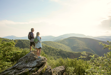 A couple on the summit of a hike surrounded by forest-covered mountains.