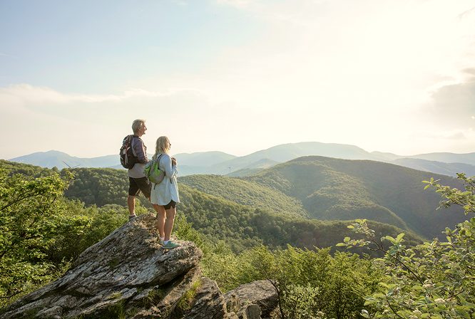 A couple on the summit of a hike surrounded by forest-covered mountains.